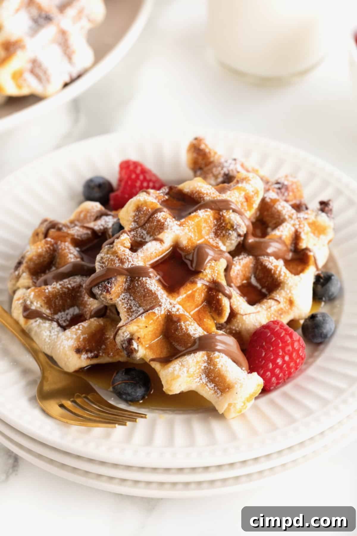 A large white fluted plate with two waffle-textured pastries topped with powdered sugar, maple syrup, a Nutella drizzle, blueberries and strawberries.