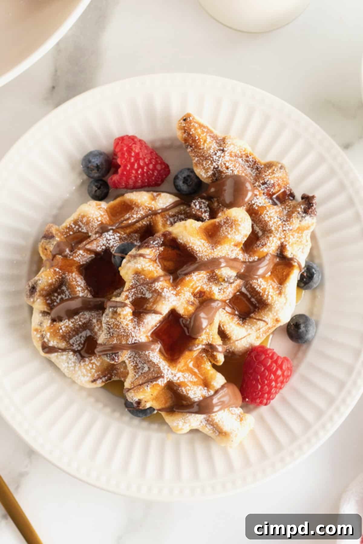 A large white fluted plate with two waffle-textured pastries topped with powdered sugar, a Nutella drizzle, maple syrup, blueberries and raspberries.