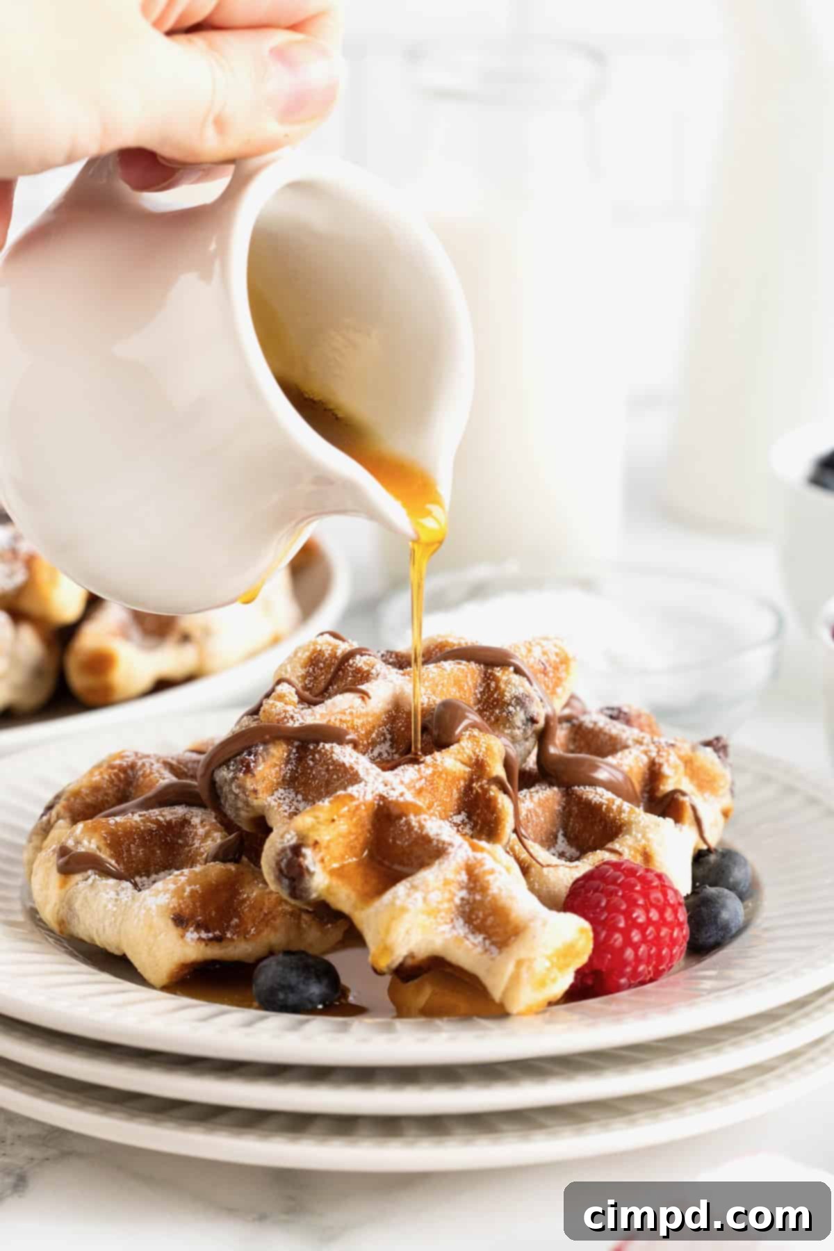 Maple syrup being poured from a small white porcelain pitcher onto two Nutella croffles on a white fluted plate.