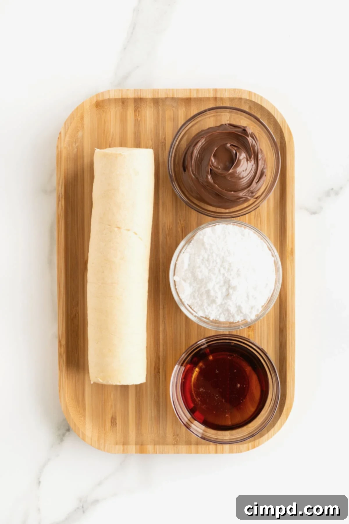 Store bought crescent roll dough and three small glass dishes of Nutella, powdered sugar and maple syrup on a rimmed wooden cutting board.