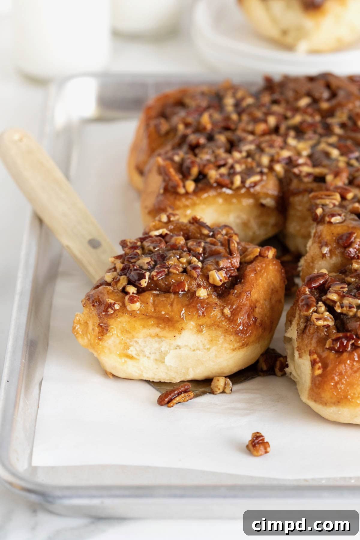 A sticky bun on a spatula resting on a parchment lined aluminum baking sheet of sticky buns.