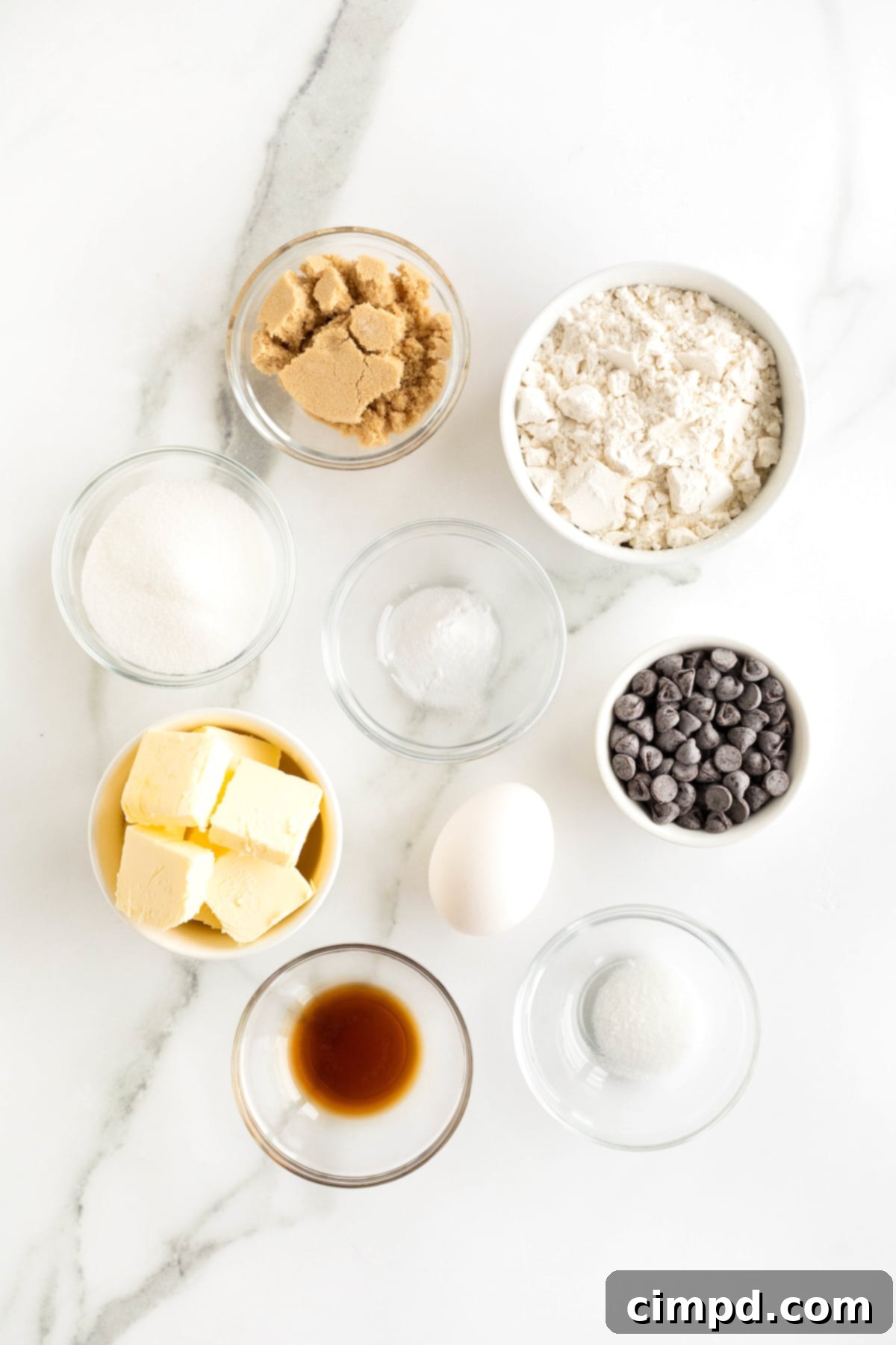 The Ultimate Chocolate Chip Cookies 6 Ingredients for classic chocolate chip cookies are neatly arranged in small glass dishes on a pristine white marble counter, ready for baking.
