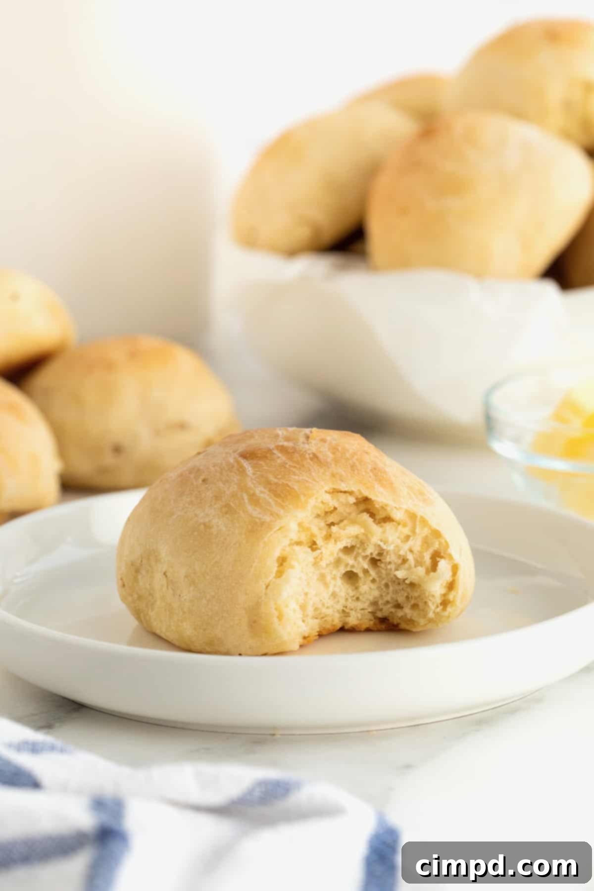 Traditional Dinner Rolls 3 A single classic dinner roll, with a small bite taken out, rests on a pristine white rimmed plate. In the softly blurred background, a bowl brimming with more dinner rolls and a small dish of butter hint at a delicious meal.
