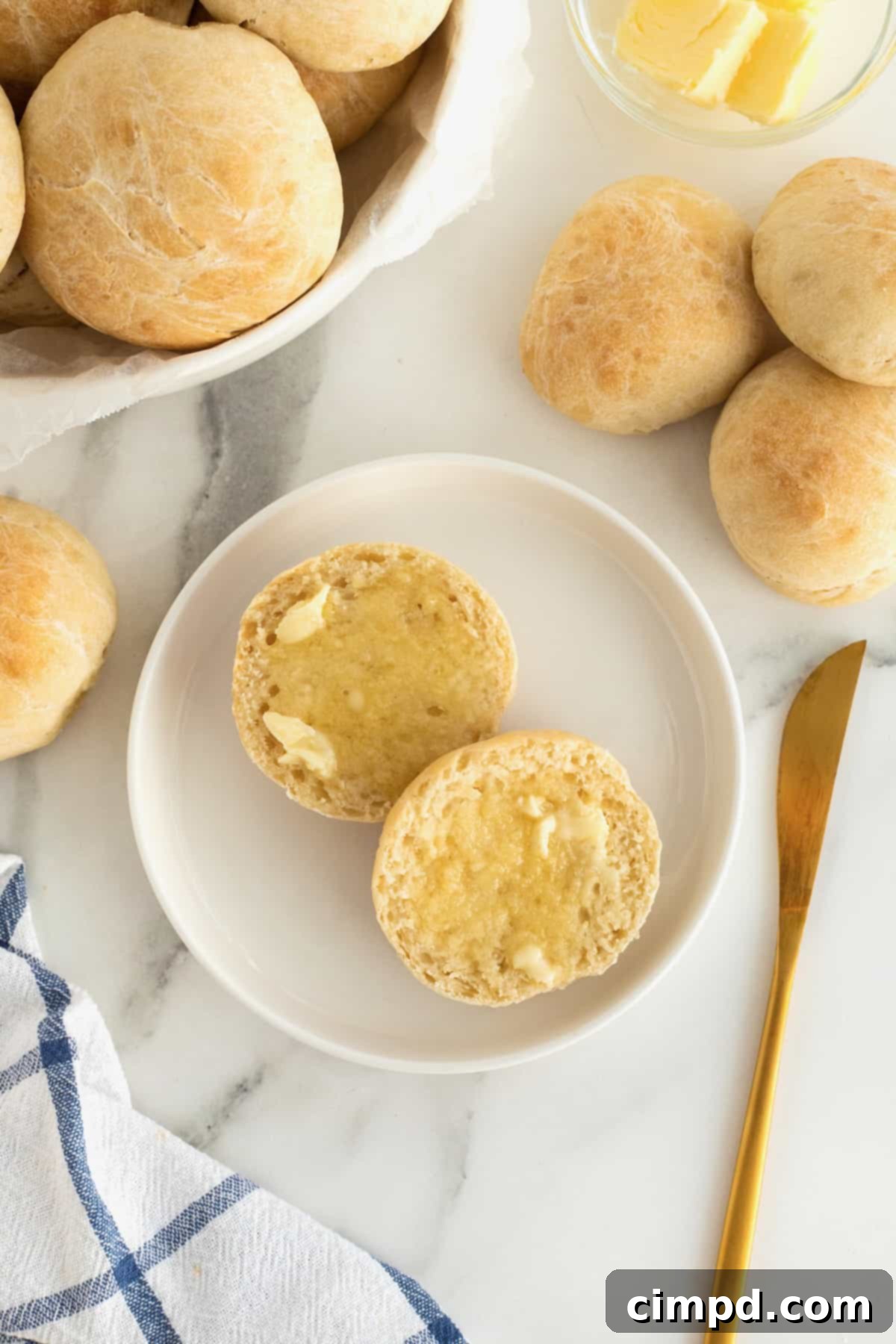 Traditional Dinner Rolls 4 A perfectly golden-brown dinner roll, cut neatly in half and generously buttered, sits on a white rimmed plate. A elegant gold-toned butter knife lies beside the plate, inviting a delicious bite.