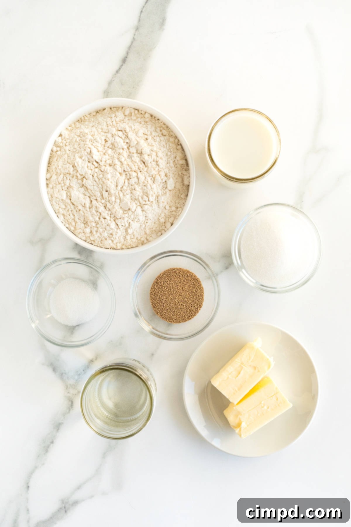 Traditional Dinner Rolls 5 Various ingredients for basic dinner rolls, including flour, sugar, yeast, and butter, neatly arranged in small glass dishes on a pristine white marble counter, ready for baking.