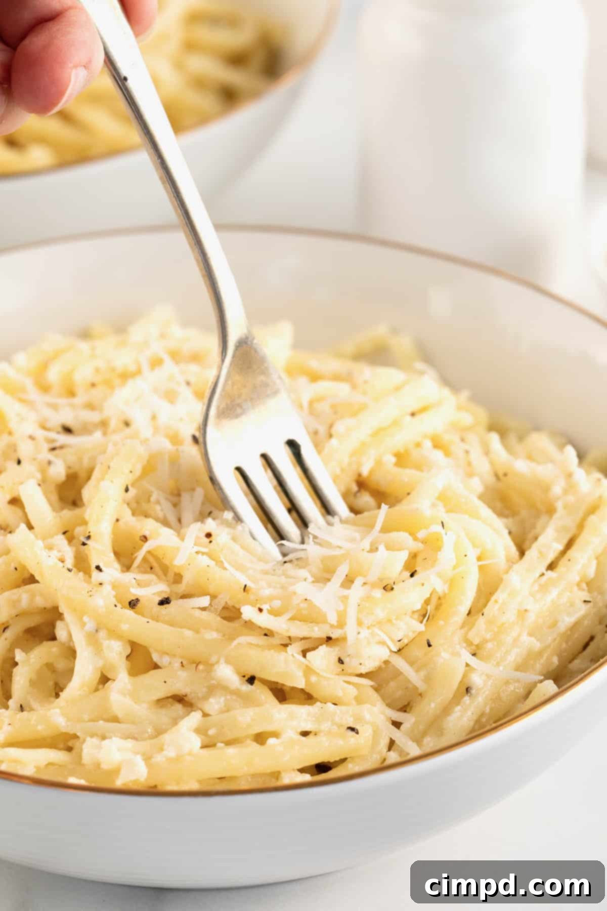 A fork twirling creamy Cacio e Pepe pasta, topped with grated Pecorino Romano cheese in a white, gold-rimmed bowl.