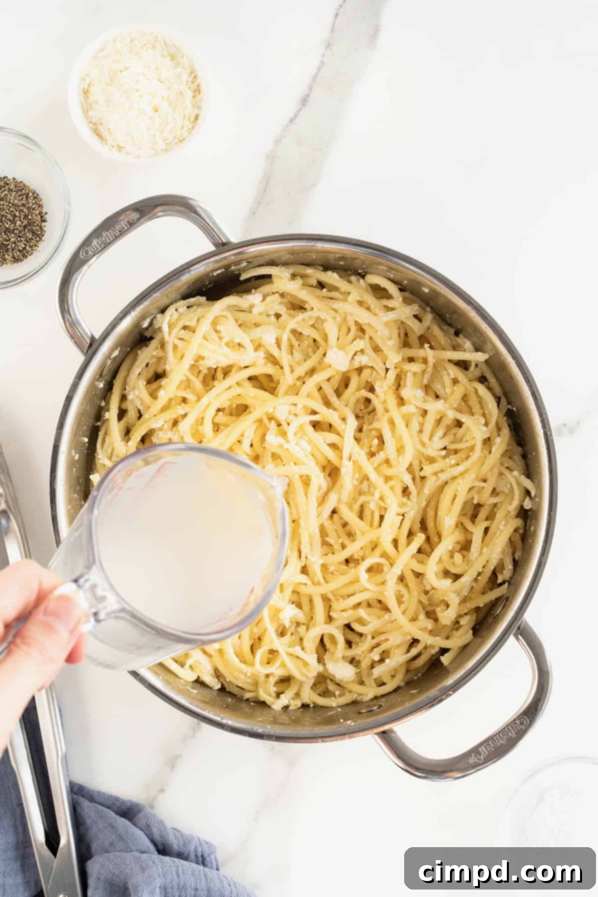 Reserved pasta water being poured from a glass measuring cup into a large stainless steel pot filled with long, thin pasta and sauce.