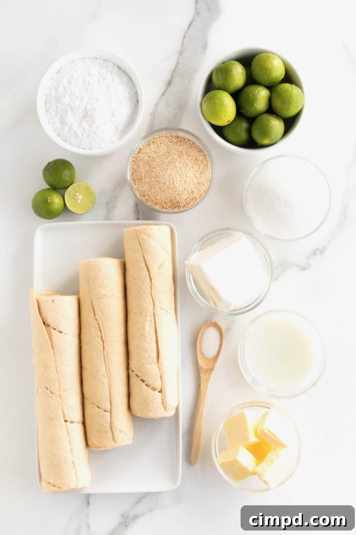 All the ingredients for Key Lime Cruffins, including crescent roll dough, butter, sugars, key limes, and cream cheese, neatly arranged in small glass containers on a white marble counter.