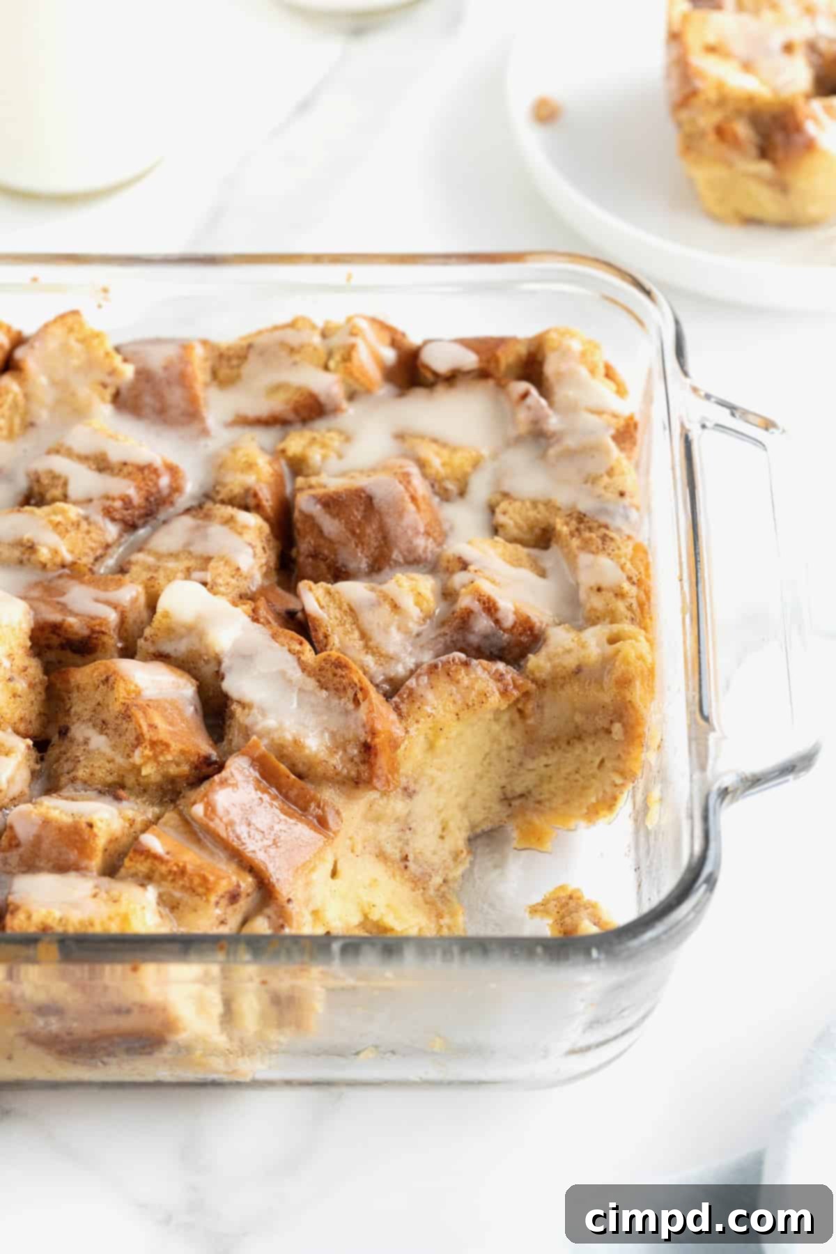 A square glass baking dish of bread pudding with white icing drizzled on top. The bottom right hand corner of the dish has the bread pudding scooped out.