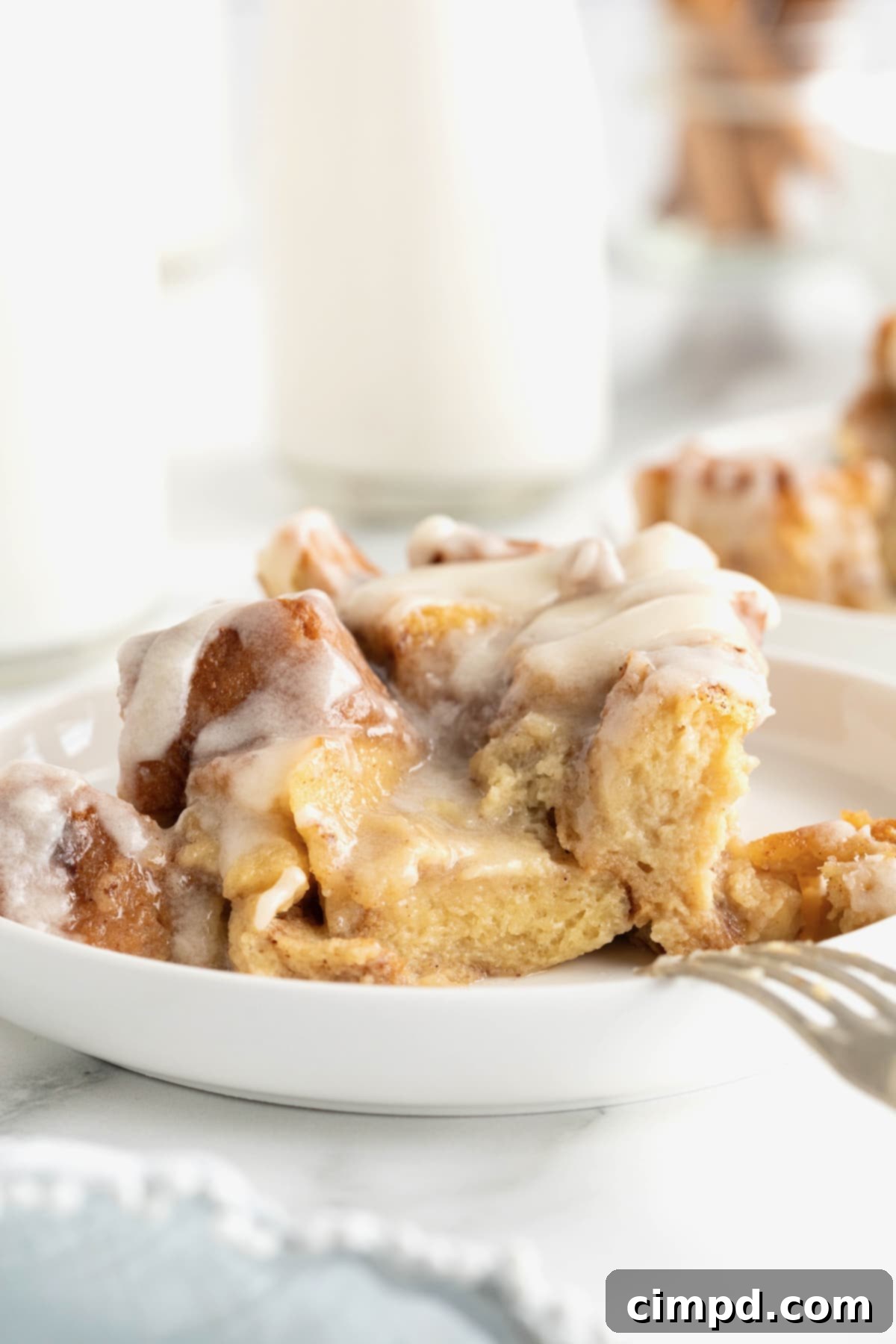 A small white rimmed dessert plate with a serving of bread pudding topped with a white sauce. There is a fork leaning on the plate.