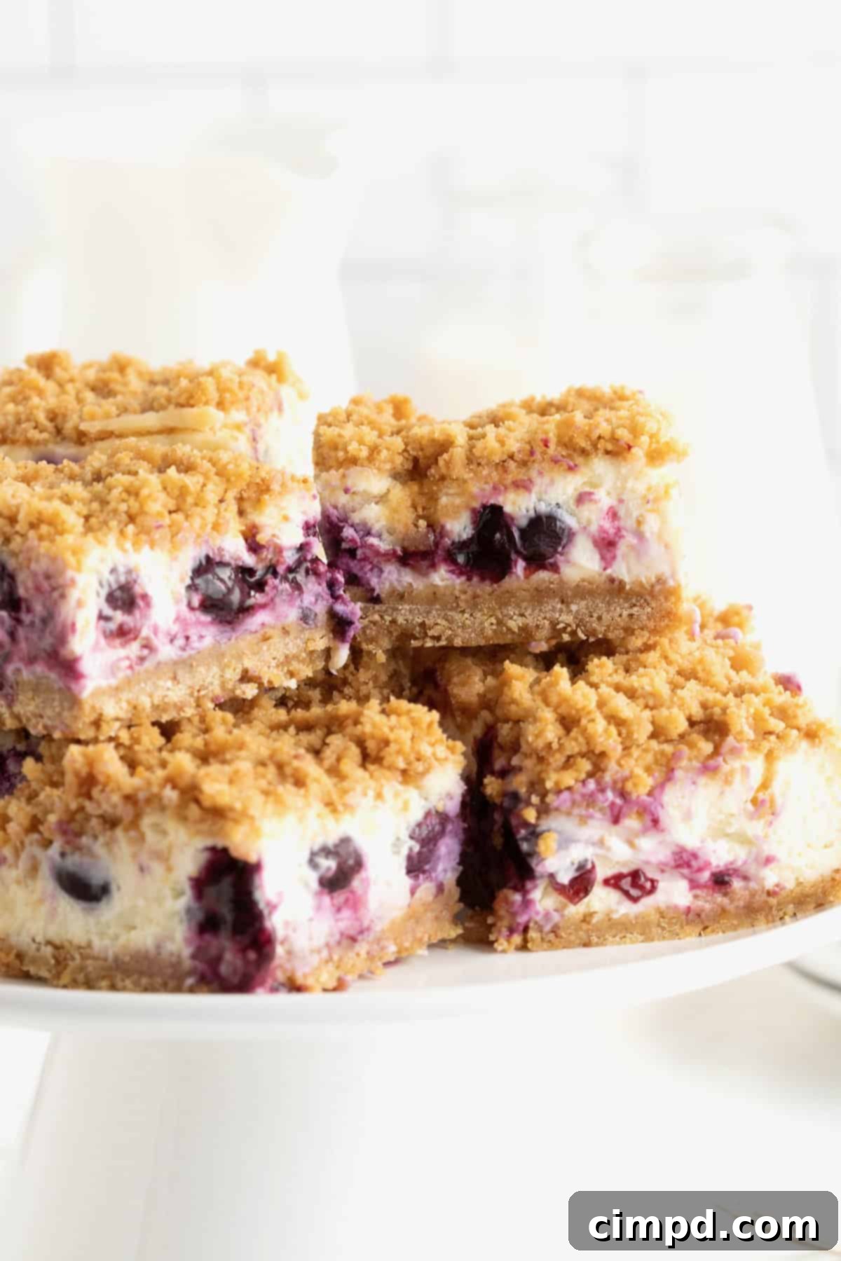 Four perfectly portioned lemon blueberry cheesecake bars on an elegant white cake stand, showcasing their golden streusel topping and vibrant blueberry filling.