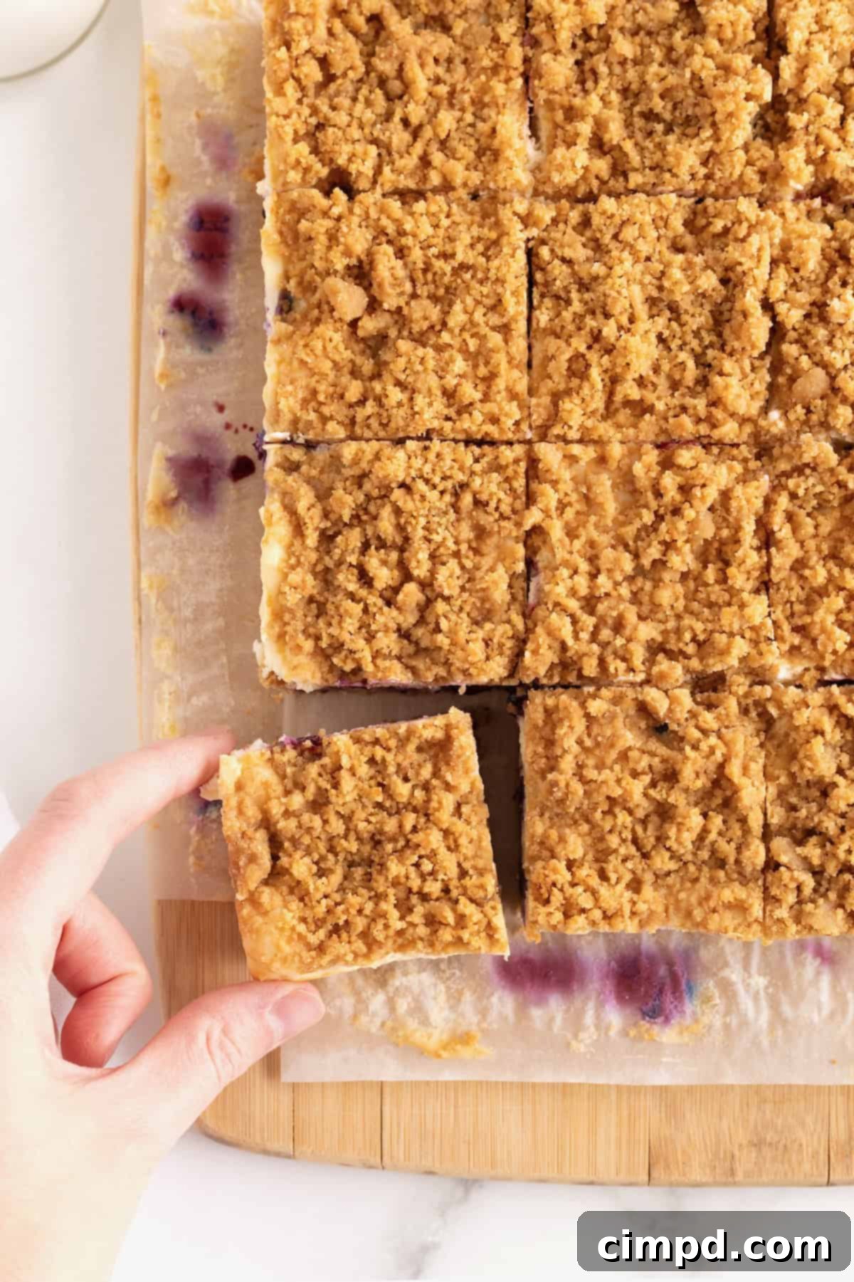 A hand reaching to pick up a lemon blueberry cheesecake bar from a group of 12 squares neatly arranged on a parchment-lined cutting board, showcasing their ready-to-eat appeal.