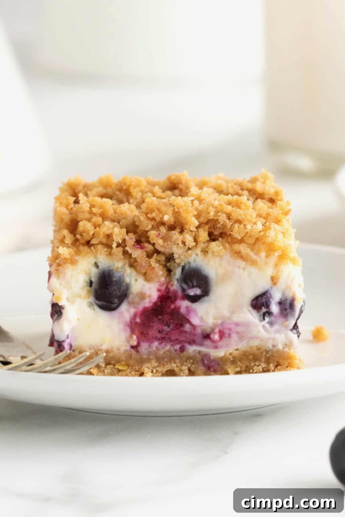 A close-up side view of a lemon blueberry cheesecake bar resting on a small white dessert plate, with a fork gently placed beside it.