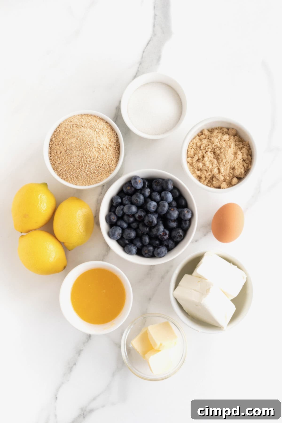 A curated display of ingredients for lemon blueberry cheesecake bars, arranged in small white dishes on a pristine white marble counter, ready for baking.