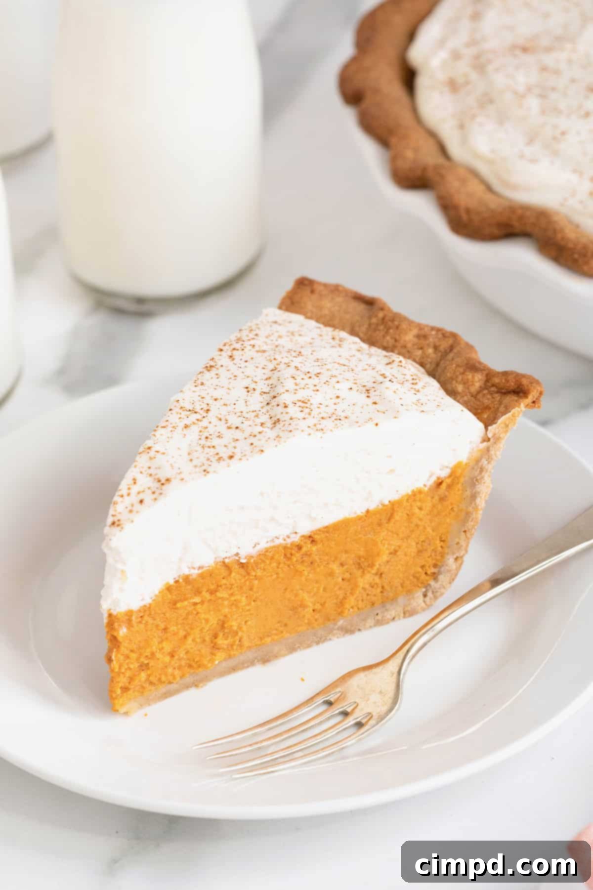 A slice of pumpkin pie with a layer of whipped topping on a white dessert plate. There is a fork next to the pie.