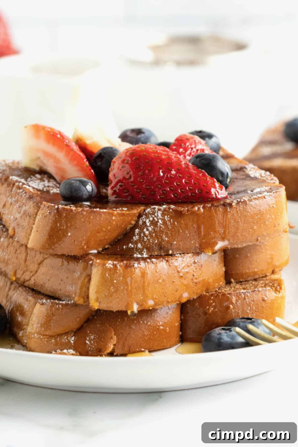 Three slices of baked French toast stacked on a white plate. The French toast is topped with blueberries, strawberry slices and dusted with powdered sugar.