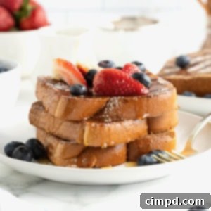 Three slices of baked French toast stacked on a white plate. The French toast is topped with blueberries, strawberry slices and dusted with powdered sugar.