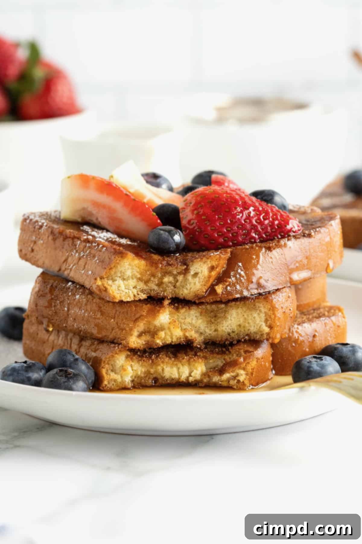 Three slices of baked French toast stacked on a white plate. The French toast is topped with blueberries, strawberry slices and dusted with powdered sugar.