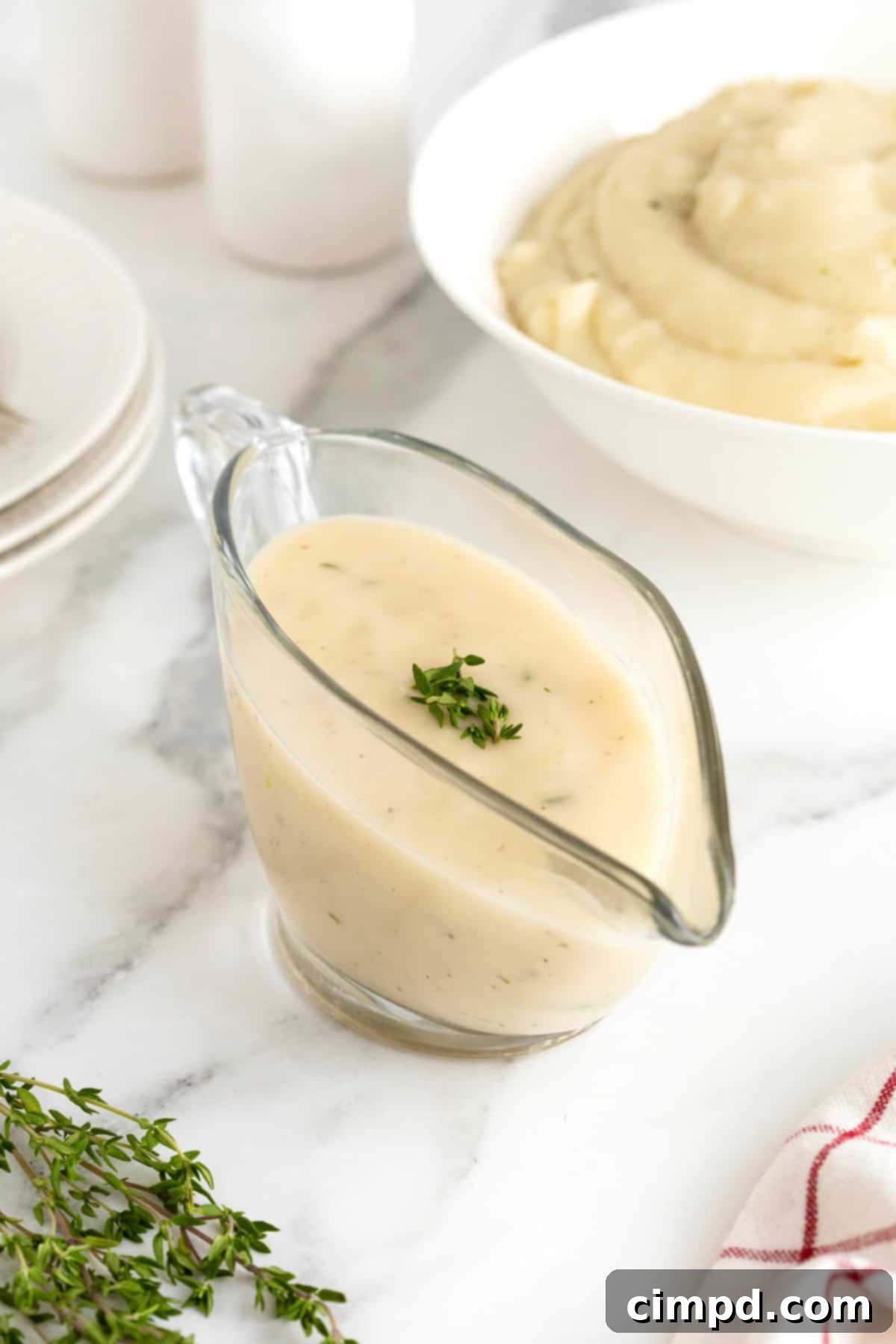 A glass gravy boat of rich, golden-brown turkey gravy on a pristine white marble counter. A large white serving bowl brimming with creamy mashed potatoes is softly blurred in the background, hinting at the perfect pairing.