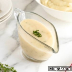 A glass gravy boat of turkey gravy on a white marble counter. A white serving bowl of mashed potatoes is in the background.
