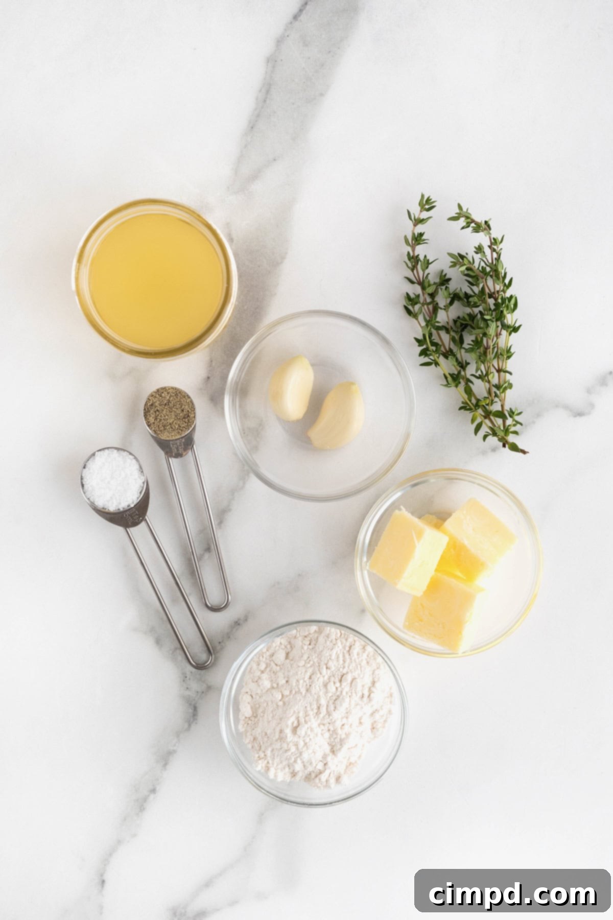 Various ingredients for turkey gravy, including butter, minced garlic, flour, stock, and fresh thyme, neatly arranged in small glass dishes on a white marble counter.