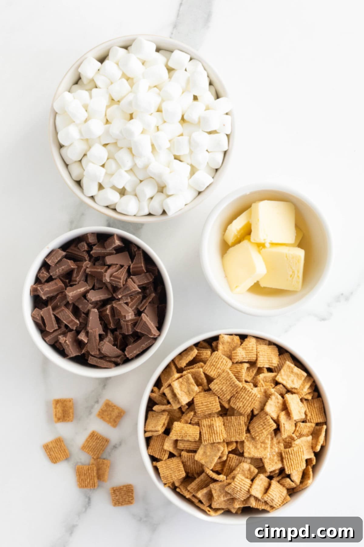 Easy No Bake Smores Bars 6 Various ingredients for No-Bake S'mores Treats, including butter, marshmallows, Golden Grahams, and Hershey's chocolate, displayed in small white glass dishes on a white marble counter.