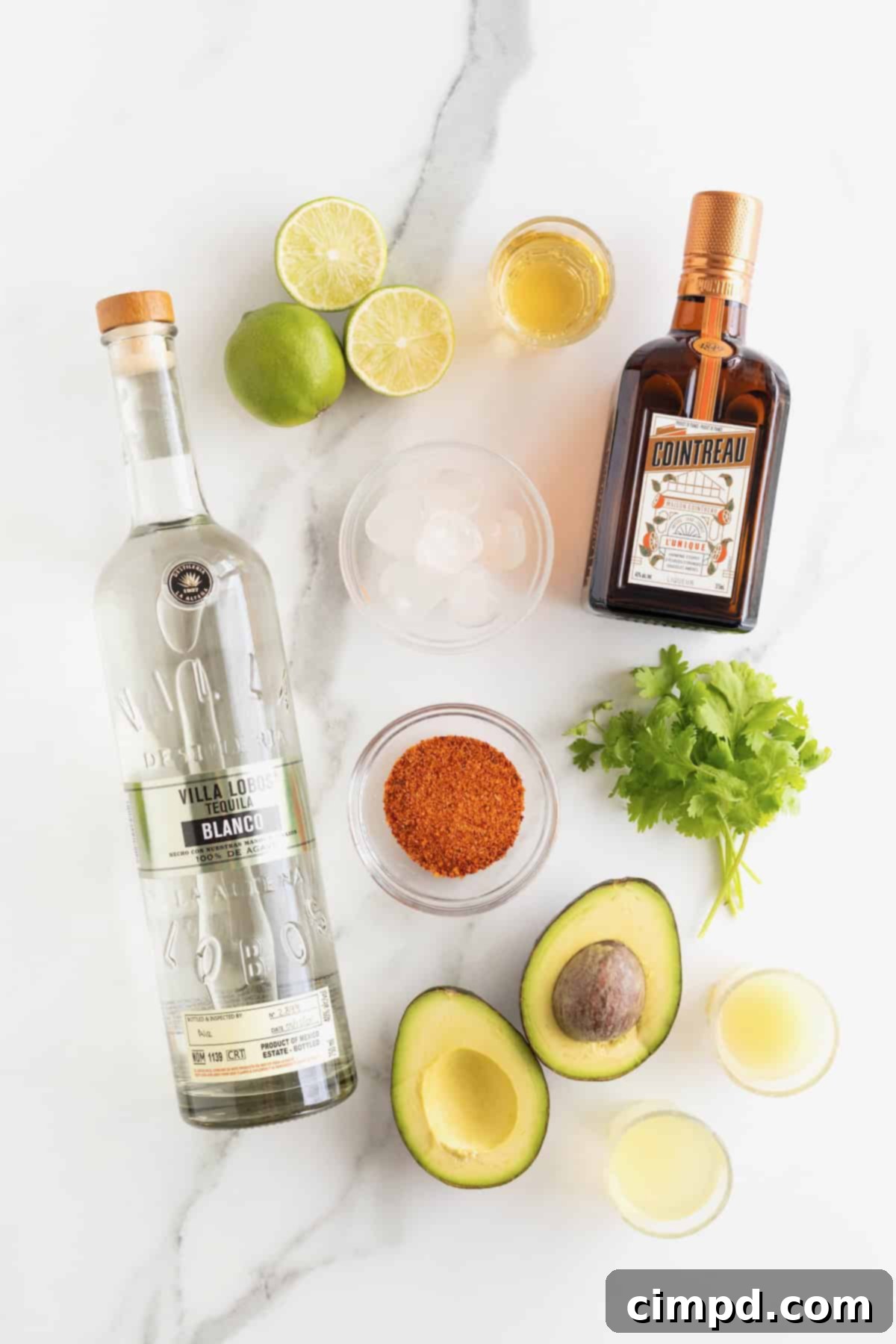 A beautifully arranged selection of fresh ingredients to make an avocado margarita, including a halved avocado, limes, fresh cilantro, bottles of tequila and orange liqueur, and a shaker, all set on a clean white marble counter.