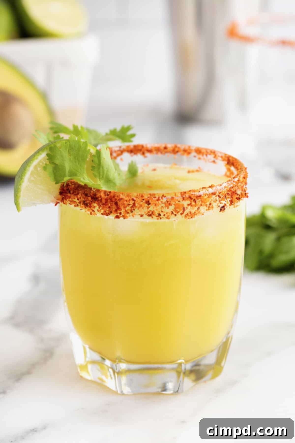 A short, clear glass brimming with a perfectly crafted avocado margarita, its rim beautifully coated with a vibrant red Tajín seasoning. The drink's refreshing green hue is visible through the glass.