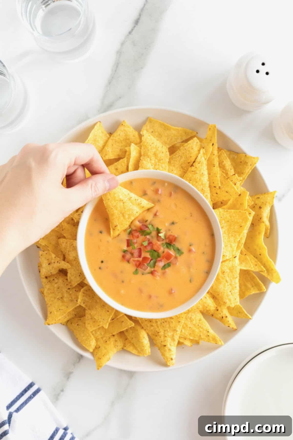 A bowl of creamy cheddar queso sits on a large white round serving platter, generously surrounded by golden corn tortilla chips, ready for dipping.