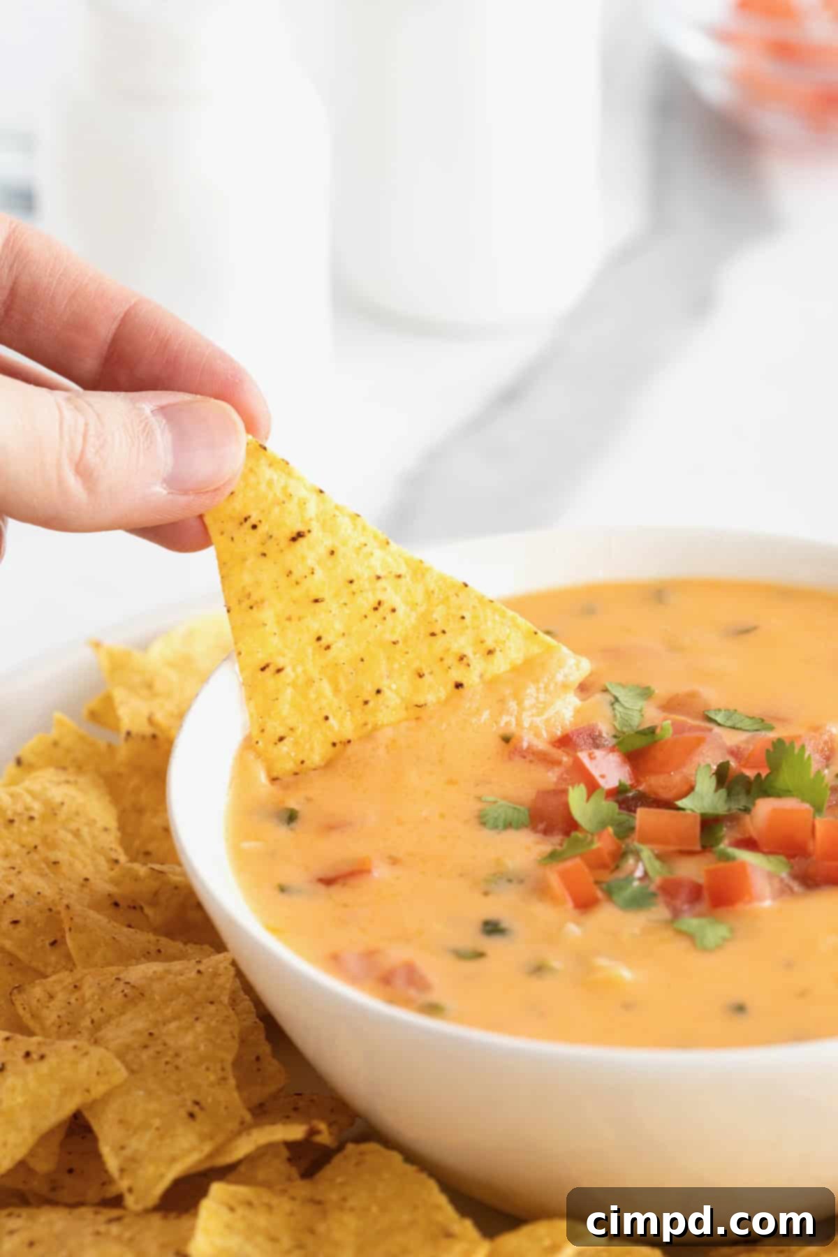 A hand gently dips a crisp corn tortilla chip into a pristine white serving bowl filled with a rich, melted queso dip, highlighting its creamy texture.