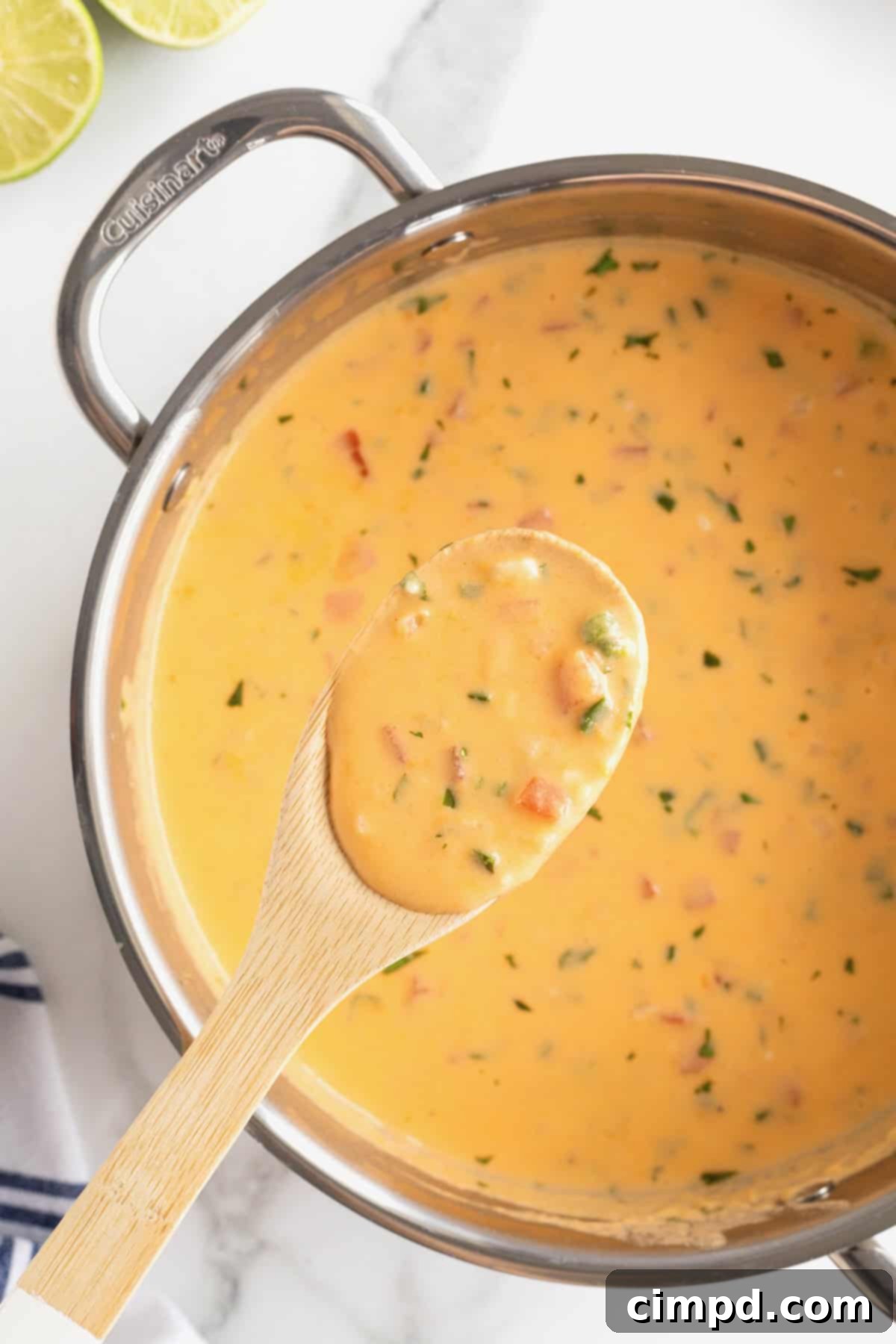 A large, gleaming stainless steel pot is filled with a bubbling, golden queso dip, with a wooden spoon holding a generous scoop of the creamy cheese hovering just above, showcasing its perfect melted consistency.