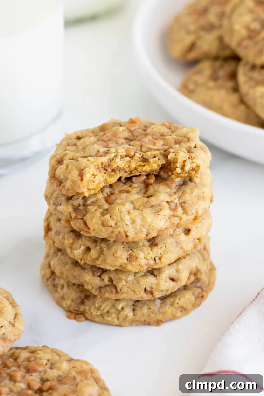A perfectly stacked pile of five chewy Brown Sugar Oatmeal Toffee Crunch Cookies on a sleek white marble counter, with the top cookie showing a delightful bite taken out, inviting a taste.