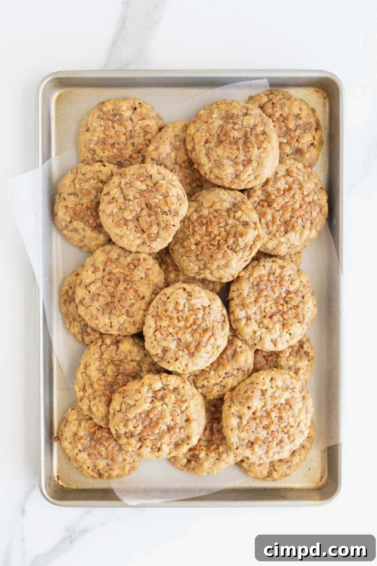 A parchment-lined baking sheet generously piled with golden-brown Brown Sugar Oatmeal Toffee Crunch Cookies, fresh from the oven and ready to be cooled.