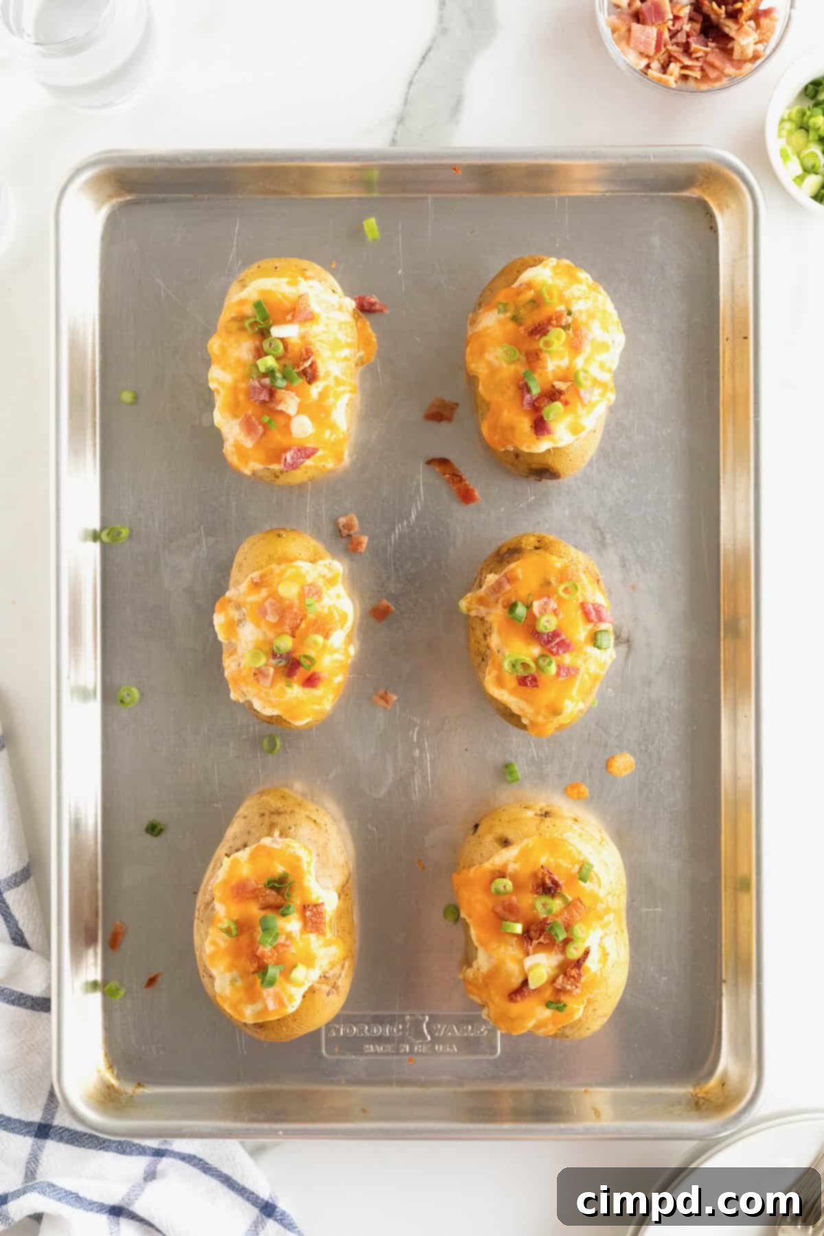 Six perfectly baked twice baked potatoes on an aluminum baking sheet, beautifully garnished with melted cheddar cheese, vibrant green onion, and crispy bacon bits, ready to be served.