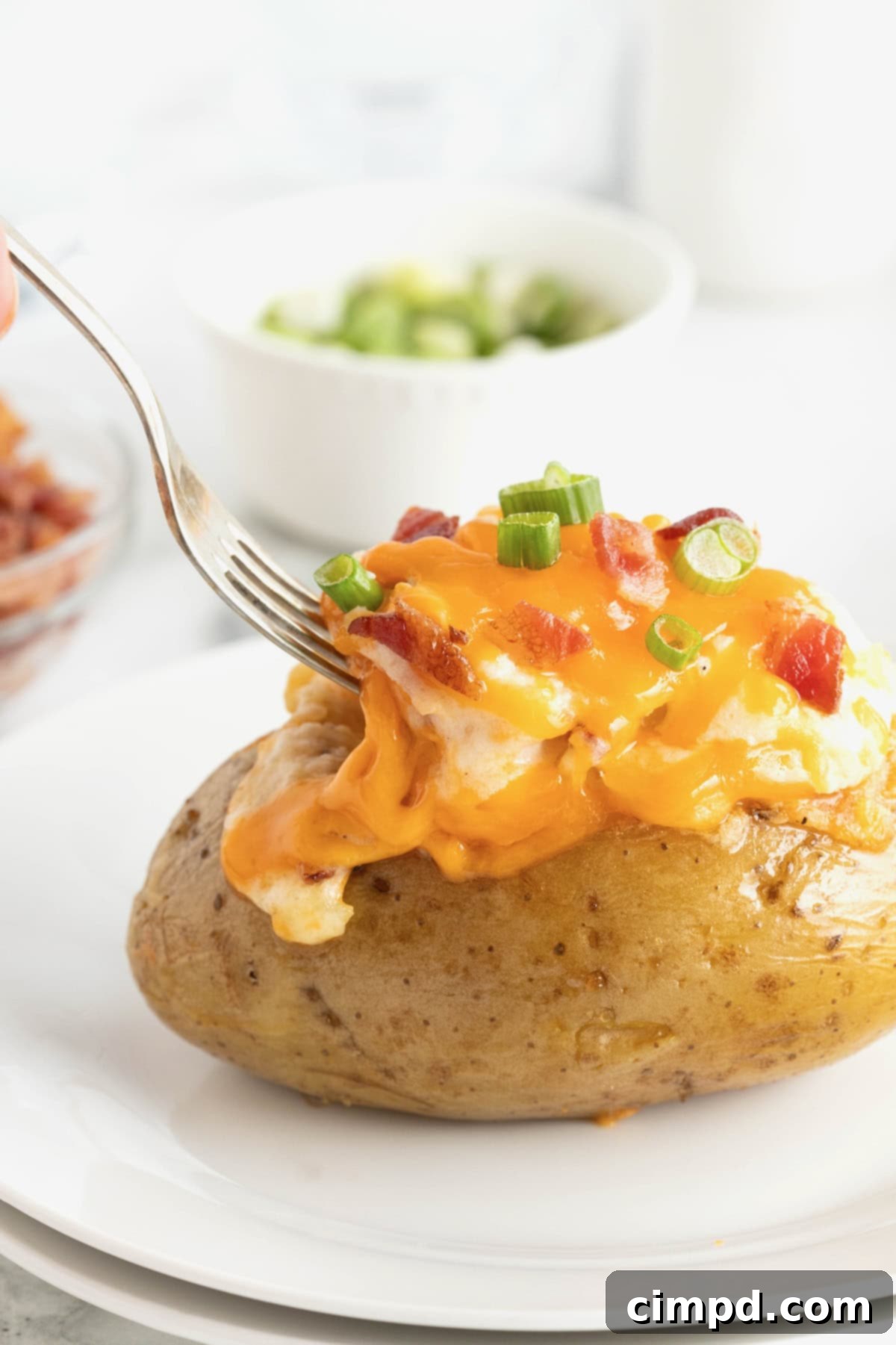A metal fork poised to dig into a perfectly crafted twice baked potato, resting on a elegant white rimmed plate. In the softly blurred background, a small dish of freshly sliced green onion awaits as a garnish.
