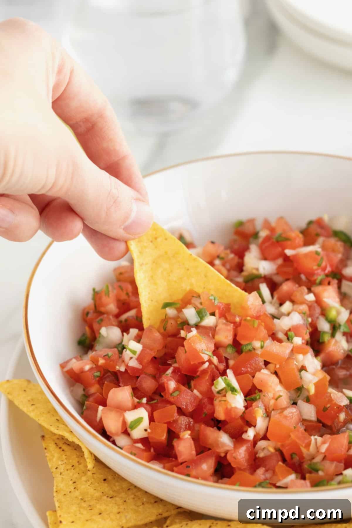 A yellow corn tortilla chip being dipped into a large white serving bowl of pico de gallo.