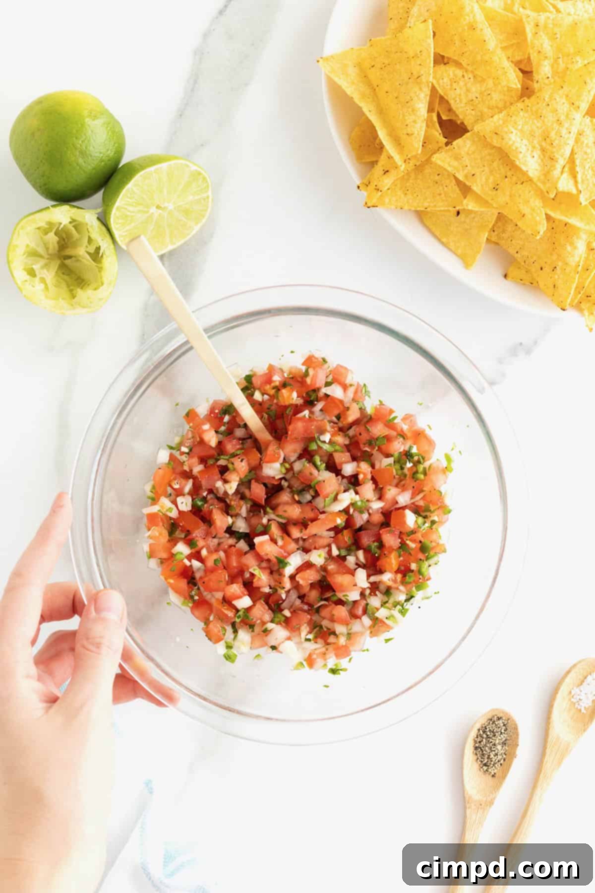 A hand holds the bottom left side of a clear glass mixing bowl of pico de gallo. In the upper right of the photo is a bowl of tortilla chips.  