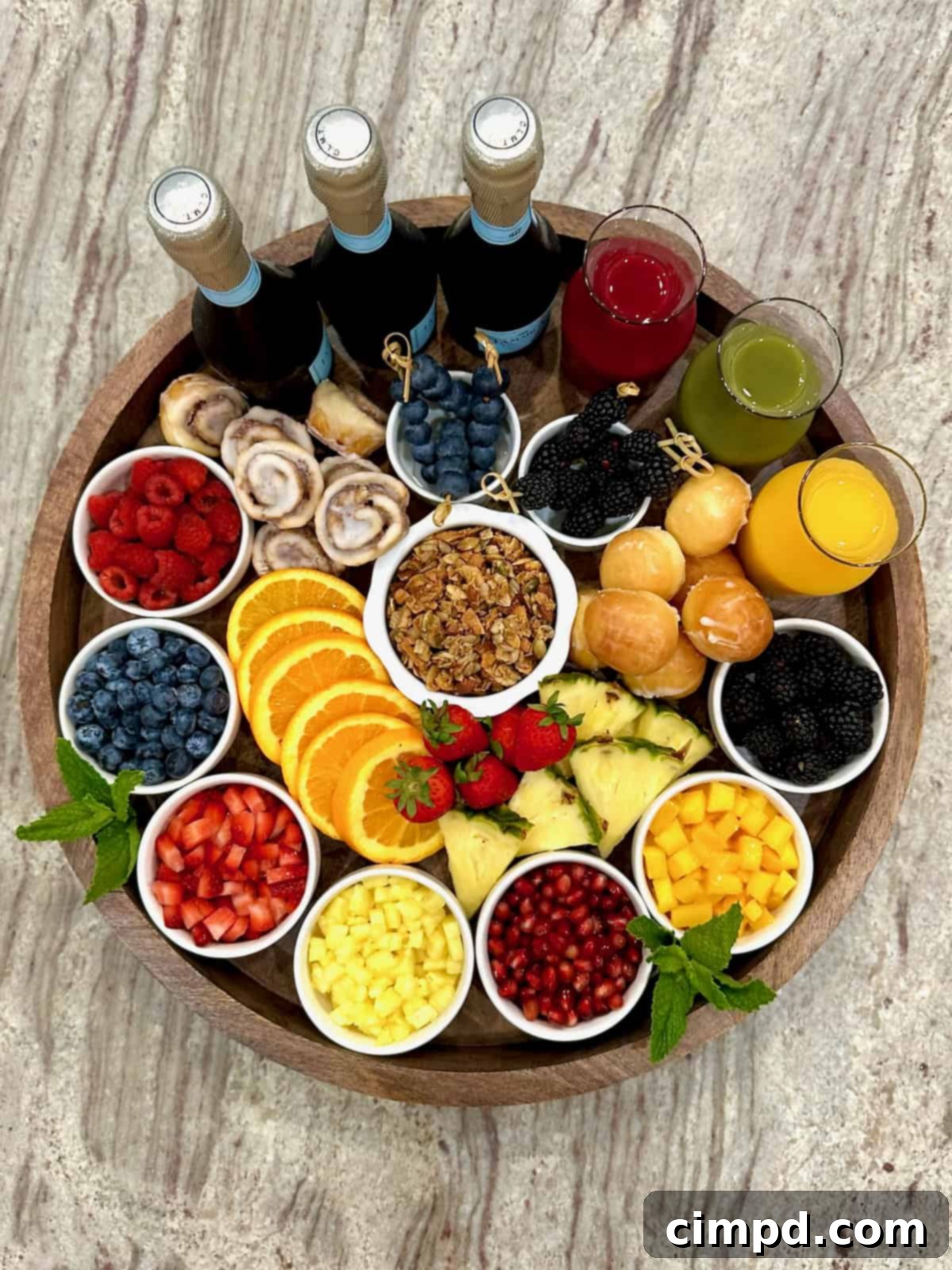 Mimosa Blossom Board 2 A beautifully arranged large round, rimmed wooden food board overflowing with an assortment of colorful fresh fruits in small white dishes, elegant glass carafes filled with various fruit juices, and three bottles of Prosecco, ready for a mimosa bar. Scattered around the board are enticing mini cinnamon rolls, light and airy donut holes, and a bowl of crunchy granola.