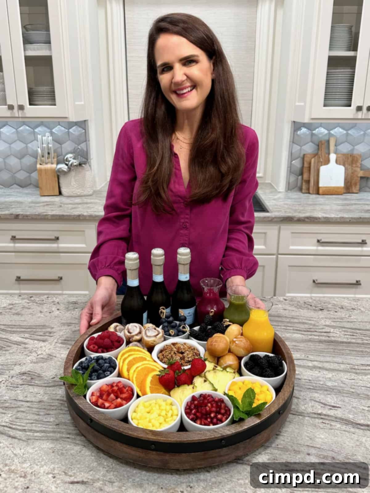 Mimosa Blossom Board 3 A radiant woman with long, dark hair, dressed in a vibrant magenta long-sleeve top, stands smiling behind a beautifully arranged mimosa board, gesturing towards the inviting spread of juices and fruits.