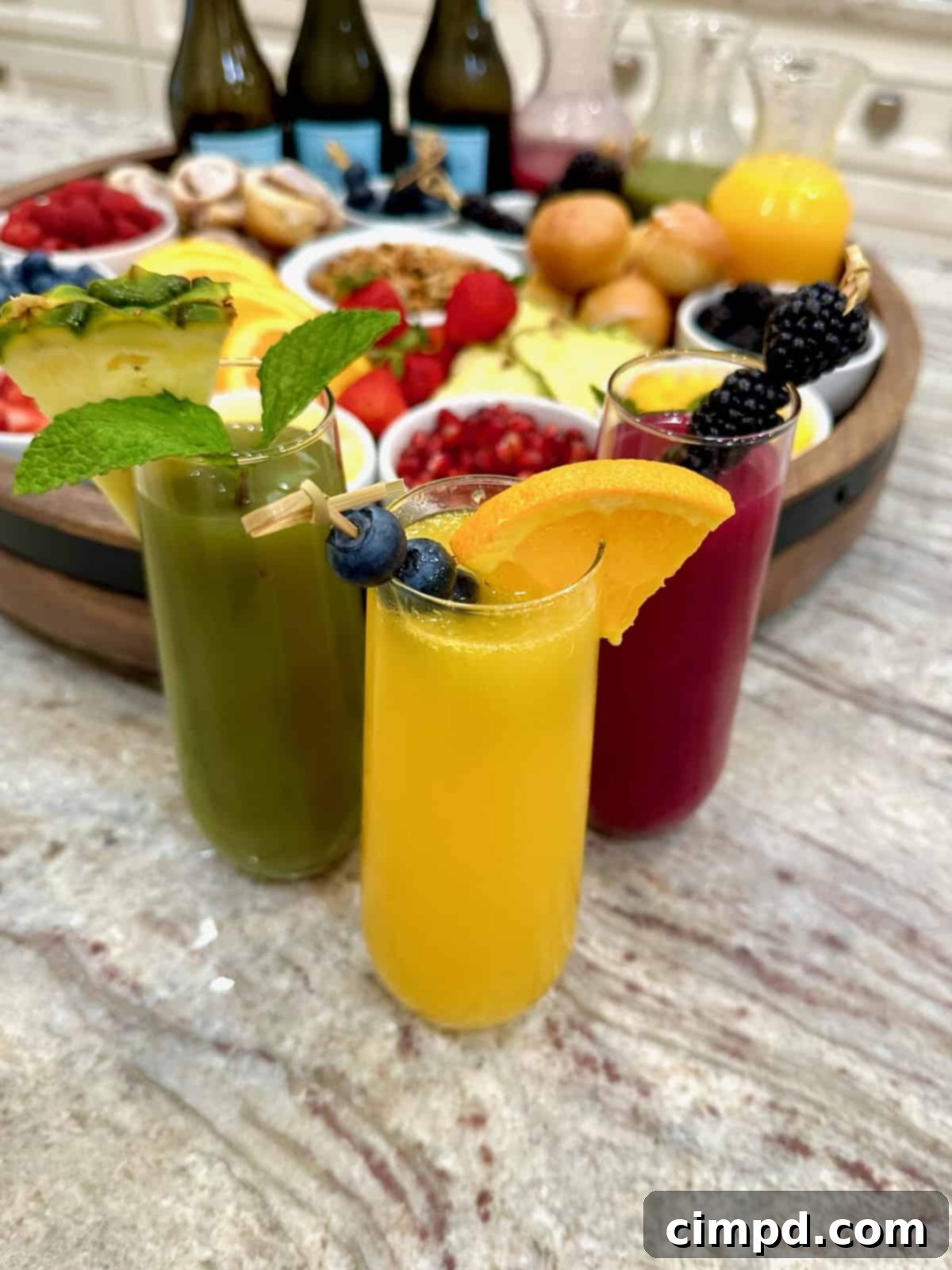 Mimosa Blossom Board 4 Three colorful mimosa cocktails, featuring vibrant hues of green, orange, and red, are beautifully presented on a sleek marble counter. Behind them, a large, round, rimmed wooden food board acts as a backdrop, filled with an inviting array of fresh fruits in small white dishes, elegant glass carafes of fruit juice, and bottles of Prosecco, suggesting a lively brunch gathering.