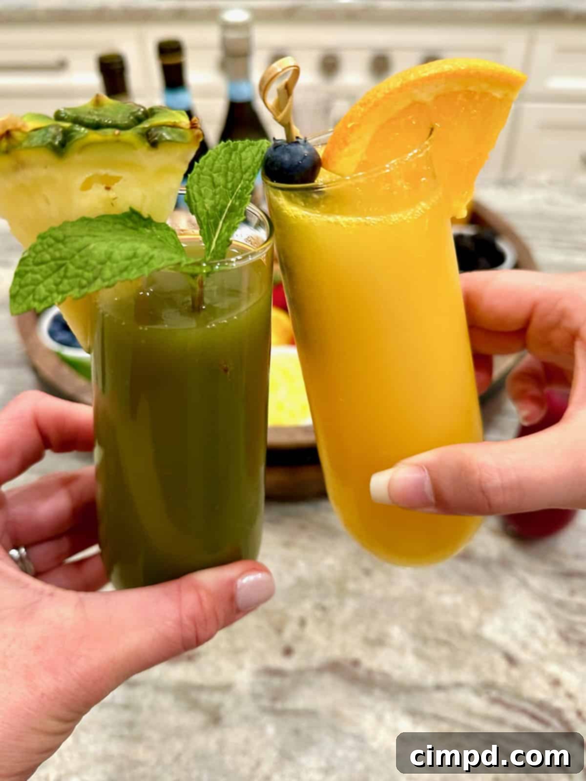 Mimosa Blossom Board 5 A vibrant green mimosa and a classic orange mimosa, both presented in elegant champagne flutes, stand in front of a beautifully arranged mimosa board. The board features an assortment of fresh fruits, various juices in carafes, and bottles of bubbly, inviting guests to create their own personalized drinks.