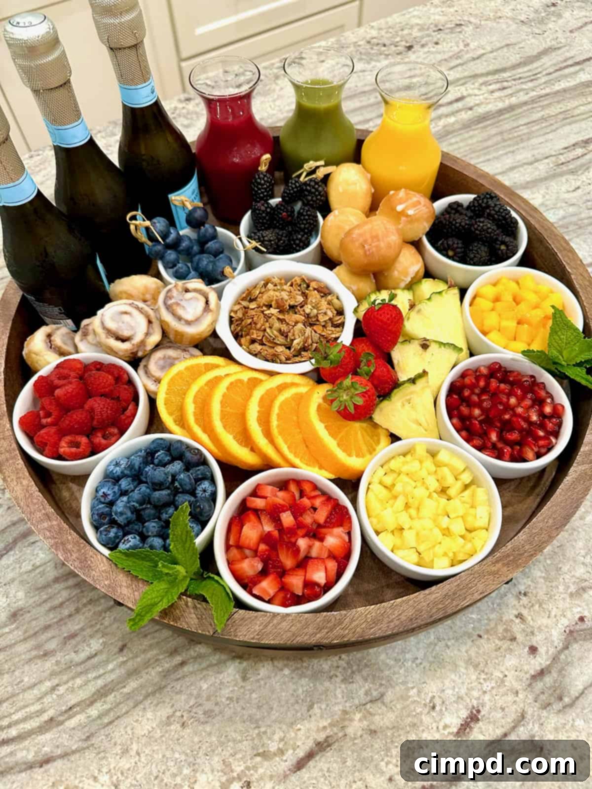 Mimosa Blossom Board 6 A large, beautifully arranged rimmed wooden Lazy Susan forms the centerpiece of a mimosa bar. It is laden with an inviting spread of colorful fresh fruits displayed in small white dishes, elegant glass carafes filled with a variety of fruit juices, and three bottles of chilled Prosecco, ready for pouring.