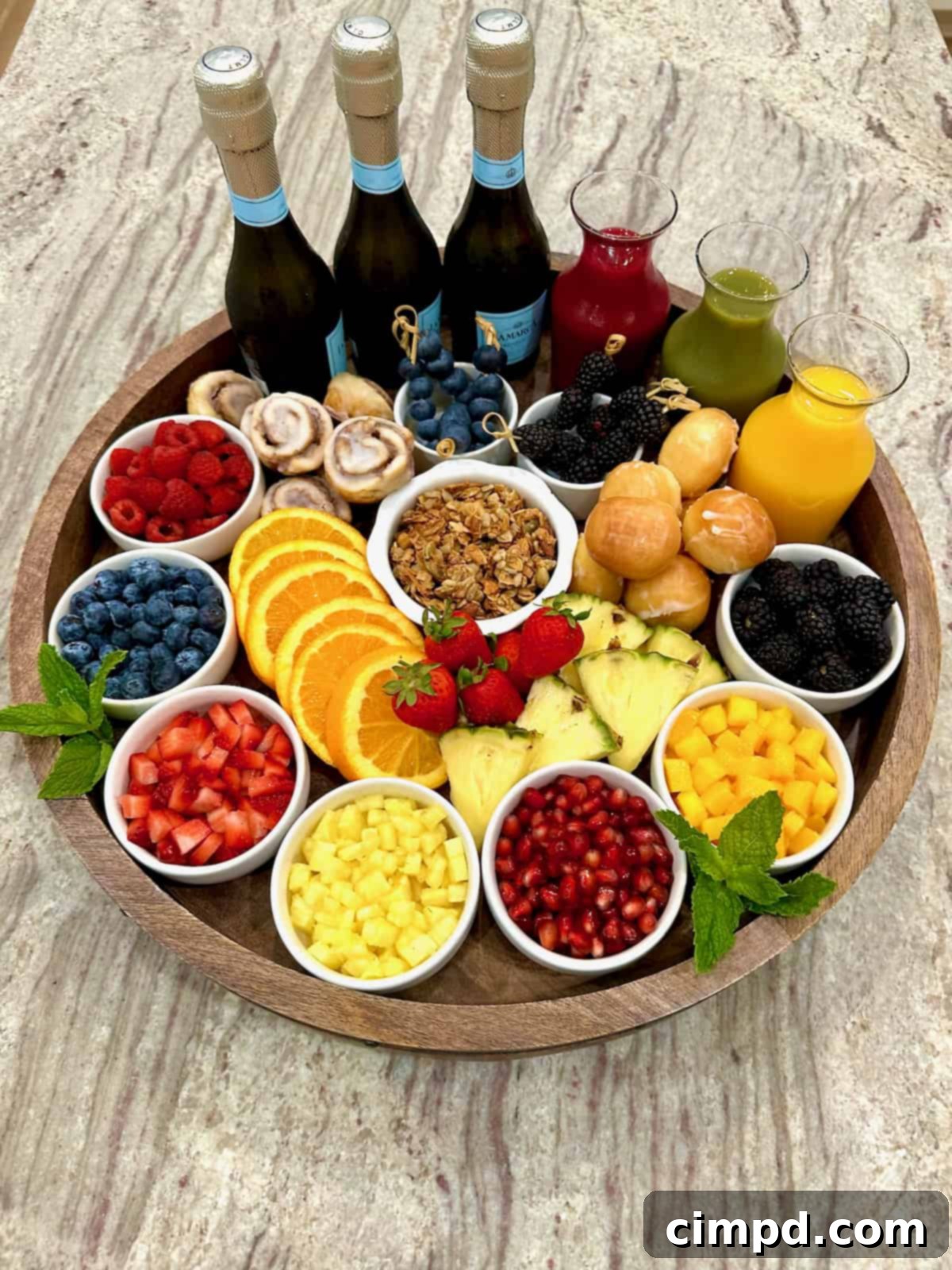 Mimosa Blossom Board 7 A large, round, rimmed wooden food board, meticulously arranged and brimming with a festive spread. It features a variety of colorful fresh fruits presented in small white dishes, elegant glass carafes filled with different fruit juices, and three bottles of Prosecco. The board is also adorned with sections of mini cinnamon rolls, delicate donut holes, and a bowl of granola, creating a perfect mimosa bar setup.