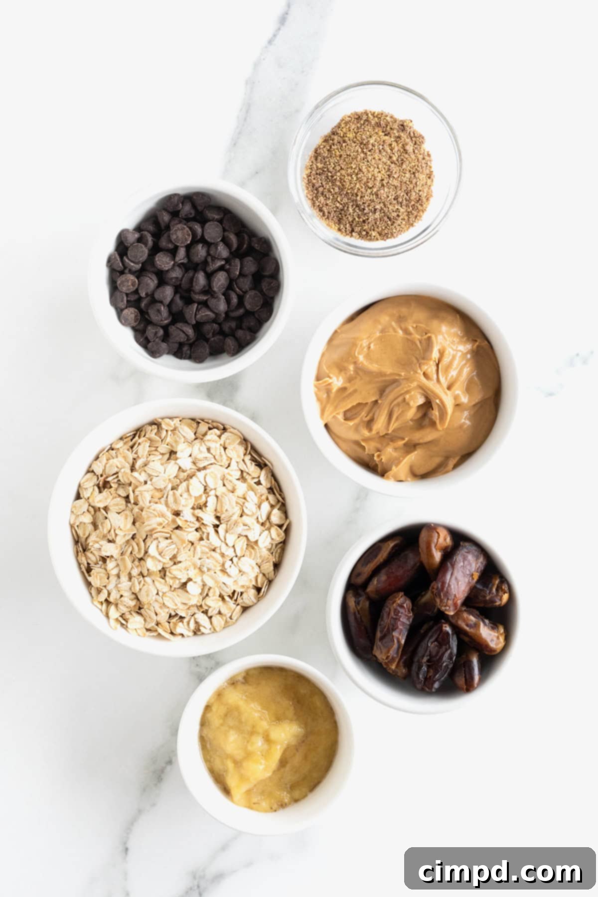 Ingredients for peanut butter chocolate chip breakfast bars with dates and banana arranged in small white glass dishes on a white marble counter, ready for preparation.