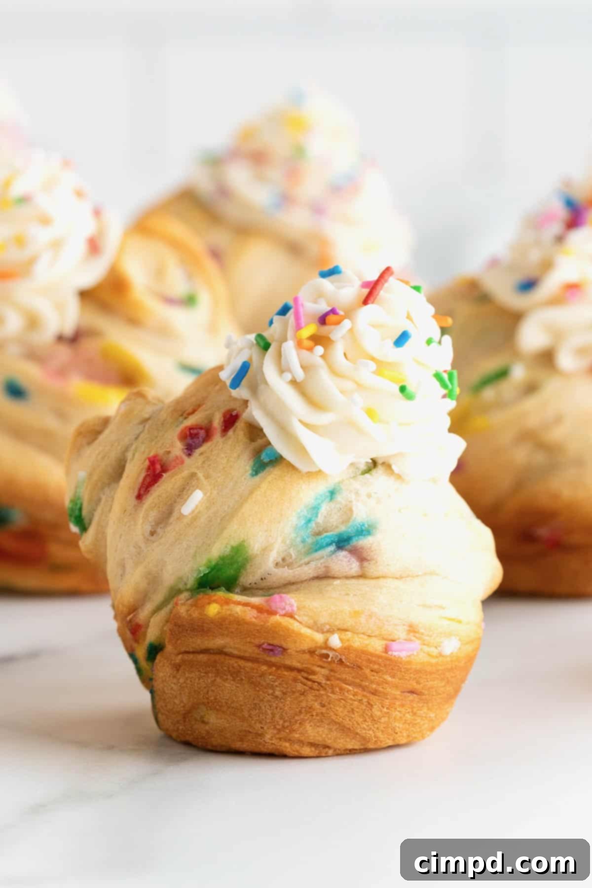 Rainbow Swirl Puffs 2 Vibrant rainbow sprinkled cruffins, generously topped with smooth vanilla icing, elegantly arranged on a pristine white marble counter, inviting a delightful indulgence.