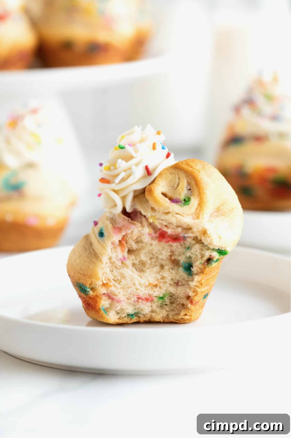 Rainbow Swirl Puffs 3 A close-up view of rainbow sprinkled cruffins, each crowned with a dollop of vanilla icing, resting on a white marble counter. One cruffin shows a delightful bite taken out, revealing its fluffy, sprinkle-filled interior.