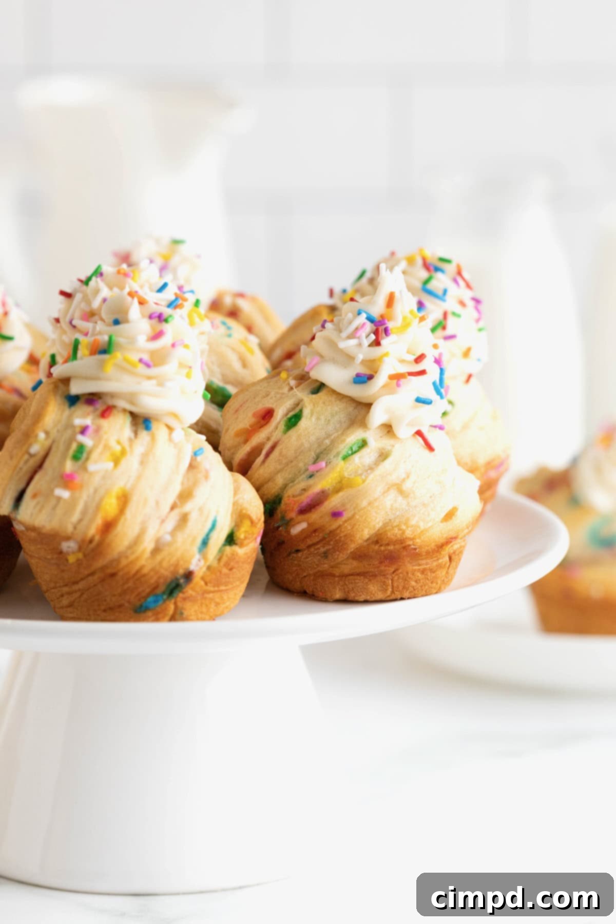Rainbow Swirl Puffs 4 A charming white cake stand showcases five beautifully frosted birthday confetti cruffins, artfully captured from a side angle, emphasizing their festive appeal.