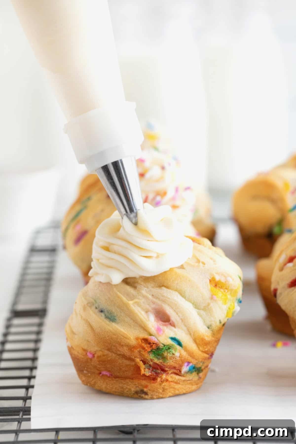 Rainbow Swirl Puffs 9 A pastry bag skillfully piping smooth white frosting onto a freshly baked confetti cruffin, ready for its final festive touch.
