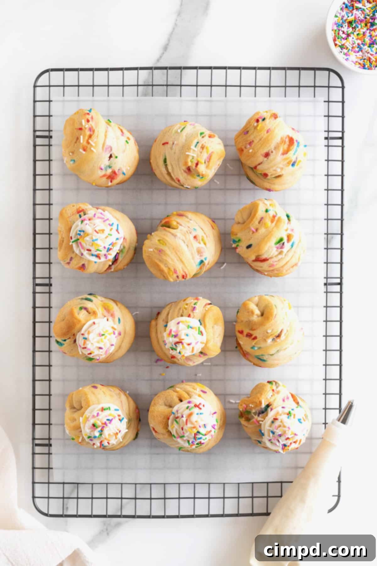 Rainbow Swirl Puffs 10 A dozen perfectly baked cruffins, each beautifully topped with vanilla icing and a shower of rainbow sprinkles, arranged on a parchment-lined metal cooling rack.