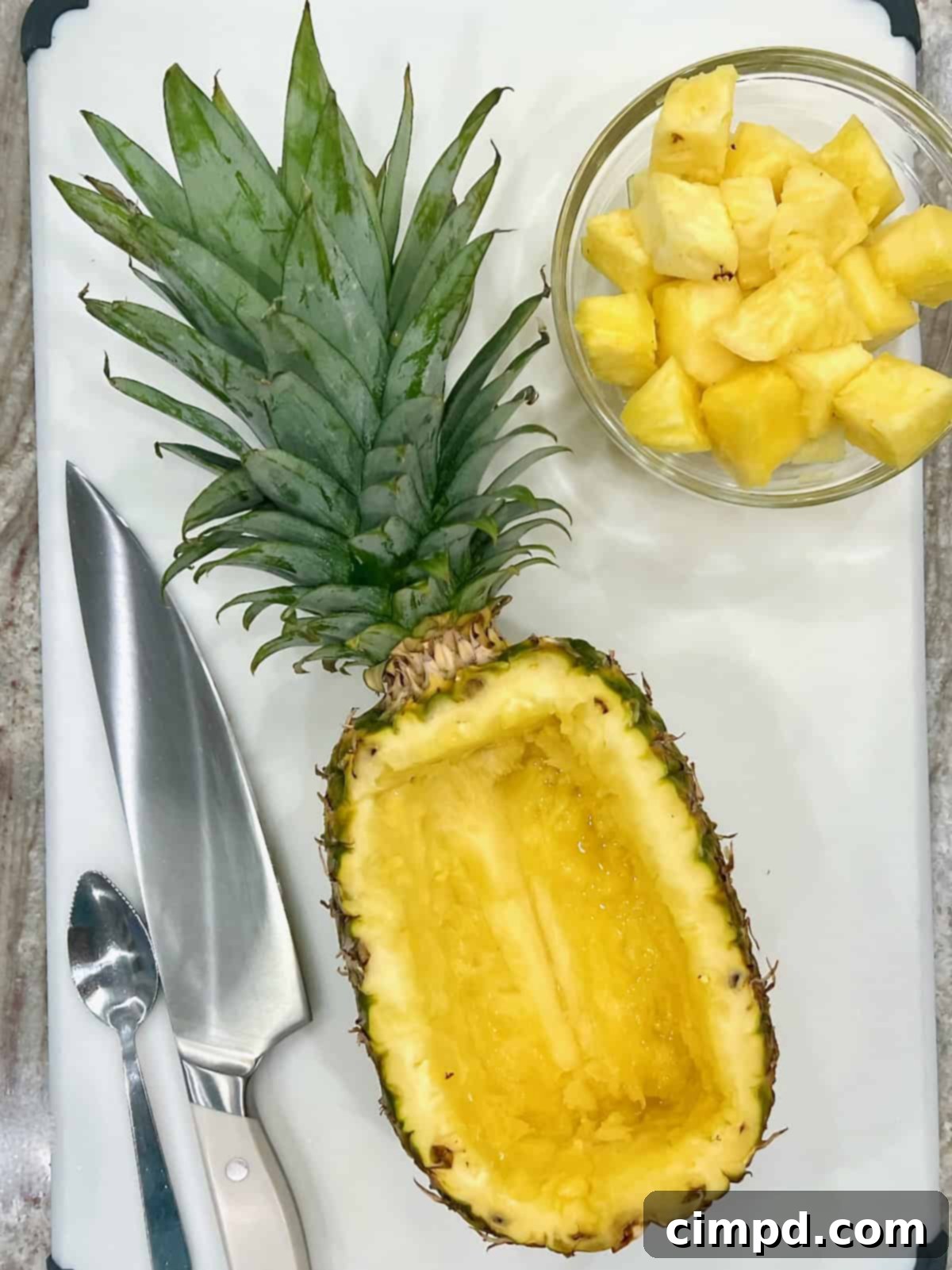 A hollowed-out pineapple half resting on a cutting board, accompanied by a glass bowl of fresh pineapple chunks and a sharp knife, ready for filling.